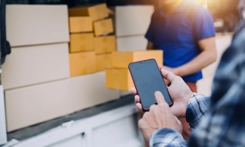 Pessoa usando smartphone para controlar estoque de celulares por IMEI ao lado de entregador com caixas em van de entrega