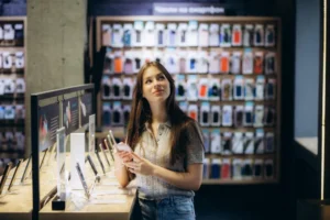 Jovem segurando smartphone em loja de celular com vitrine de aparelhos e parede de capinhas coloridas ao fundo