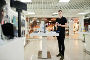 Vendedor de loja de celular segurando smartphone em corredor de shopping com mesas de exposicao de aparelhos ao fundo