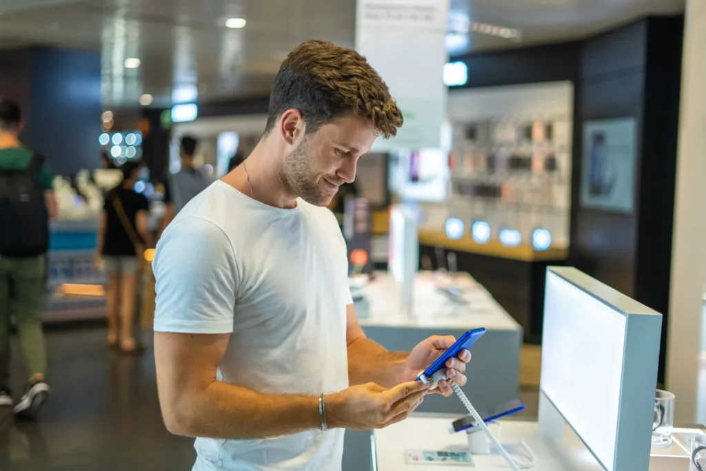Homem testando smartphone azul na vitrine de loja de celular com sistema de gestao para controle de estoque e vendas ao fundo