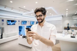 Homem de oculos e camisa rosa sorrindo ao usar smartphone em loja de tecnologia com televisores e produtos ao fundo